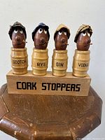 Front view of four carved wood English yokel cork stoppers on a labeled wooden display stand.