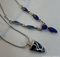Two vintage sterling silver necklaces displayed on white surface, showing one with multiple blue stones and one with shield-shaped pendant with enamel.