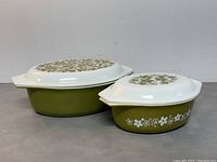 Two vintage Pyrex casseroles with lids, olive green with white floral pattern sides and lids, placed side by side on a flat surface against a plain background.