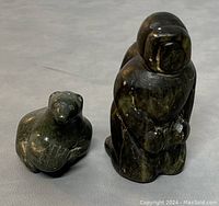 Two Inuit soapstone carvings shown on a gray surface. One is a standing human figure in traditional Inuit attire and the other is a seal figure.