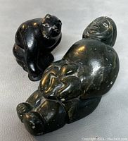 Shows two black soapstone figures on gray surface: a reclining woman holding a child and a sitting bear, highlighting size and details.