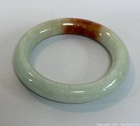 Single natural jade bangle shown on white background, angled to highlight the light green jade and red-brown area.