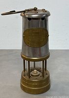 Front view of cylindrical steel body with brass base, brass nameplate, and wick visible inside.