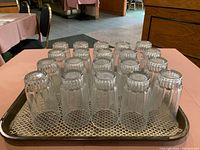 35 clear drinking glasses placed upside down inside a plastic tray with netting underneath, showing their tapered heavy bottoms and ridged design.