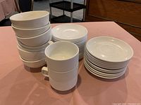 Stacked off-white bowls, cups, and two groups of saucers on a table with pink cloth.