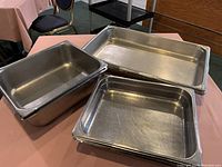 Three stacks of stainless steel buffet pans of different depths and dimensions, showing minor surface wear and scrapes.