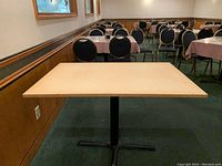Rectangular particle board table with light wood finish top and black metal base in a restaurant or meeting room with green carpet and chairs.