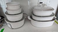 Full view of stacked white ceramic casserole and souffle dishes with metal handles and lids.