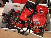 Box containing multiple Milwaukee red and black M12 chargers on right side and cordless drill with attached battery and several loose battery packs on left.