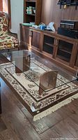 Full view of the glass top coffee table positioned on a patterned rug, showing clear rectangular glass surface and wood base with metal bar.
