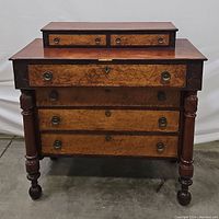 Front view of antique empire chest of drawers showing two-toned wood finish, 2 upper small drawers, 4 lower larger drawers with brass ring pulls and keyhole escutcheons, and carved round legs ending in ball feet.