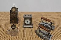 Full lot consisting of brass lantern clock, vintage metal mantel clock, finger oil lamp, and antique pair of irons, shown from front and side angles.
