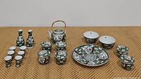 Full set of Asian ceramic tea and saki set including teapot, teacups, plate, bowls, saki bottles, and saki cups on woven mat surface.