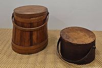 Two vintage wooden containers side by side: a barrel bucket with lid and wooden handle, and a smaller dark oval pantry box with lid and handle.