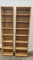 Pair of wooden bookcases shown side by side, each with 7 shelves. The left is narrower than the right.