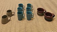Overview of all 10 mugs showing the different styles and colors: 6 teal double-handled mugs, 2 green mugs, and 2 brown stoneware mugs.