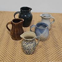 Five ceramic pitchers and vases arranged on woven surface including blue speckled cream, brown glazed, multicolored stoneware, blue-black glazed, and white with blue speckles.