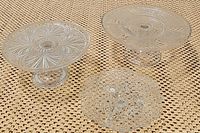 Three vintage clear glass pieces on woven fabric background: two pedestal cake stands with etched patterns and one scalloped edge footed dish.