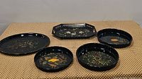 Five black hand painted metal trays arranged on beige surface; shapes include round, octagon, and oval; visible floral and basket motifs in gold, yellow, green, and cream colors.