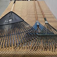 Three rakes on a perforated surface. One large plastic leaf rake with a wooden handle on the left and two metal spring back rakes with wooden handles on the right.