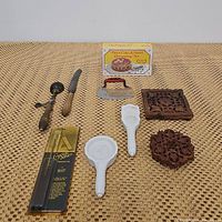 Overview of the lot showing all items arranged on a woven mat surface including two wooden trivets, two porcelain cookie molds, food chopper, ice cream scoop, spreader, decorating set and crystal saver.