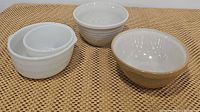 Four vintage mixing bowls on woven surface, including tan glazed ceramic, white glazed ceramic, and two white milk glass bowls.