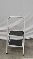 White folding 2 step stool facing front showing two black steps and white frame