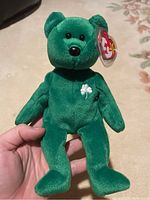 Front view of green plush Erin Beanie Baby bear with tag attached and shamrock embroidery on chest held in hand.