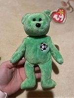 Front view of green Ty Beanie Baby bear named Kicks showing black eyes, black nose, embroidered black and white soccer ball on chest, and heart-shaped Ty tag.