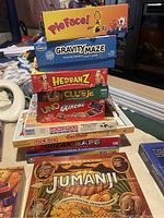 Stack of board game boxes including Pie Face, Gravity Maze, Hedbanz, Clue Jr., Uno Extreme, multiple Monopoly editions, Break Safe, and Jumanji