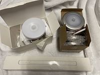 Two round LED stick-on lights in open boxes with their mounting accessories and one long LED light strip in unopened white box.