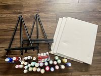 Lot contents showing three black metal tabletop easels, three blank canvases, and 24 bottles of acrylic paint in various colors.