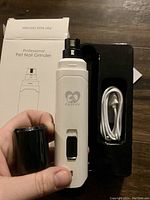 White pet nail grinder with brand name 'Casfuy', black removable cap on grinder head, and black textured power switch, held in hand with USB charging cable visible in packaging.