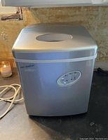 Front view of silver Salton ice maker with control panel and power cord, placed on countertop next to a candle.