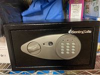 Front view of the black Sentry Safe showing electronic keypad, twist knob, and keyhole on the front panel, placed on a metal wire shelf with various items in the background.
