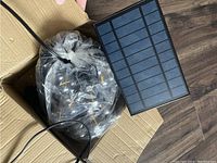 Solar powered string lights bundled in plastic inside a cardboard box, showing the string of small bulbs and solar panel with wiring.