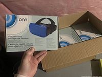 Hand holding one unopened box of Onn virtual reality smartphone headset showing blue headset image on front.