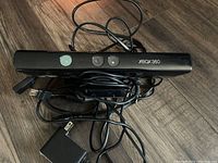 Front view of black Xbox 360 Kinect sensor bar showing three sensor lenses, Xbox 360 logo, with connected cables including power adapter and USB plug laid out on wooden surface.