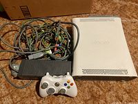 White Xbox 360 console, controller, and assorted cables on a brown carpet with a cardboard box in the background.