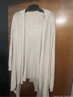Cream long open cardigan sweater with hood by Victoria's Secret, hanging on a black hanger in front of a wooden door.