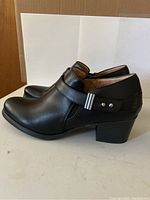 Side view of black leather ankle boot showing decorative strap with metal buckle and block heel.