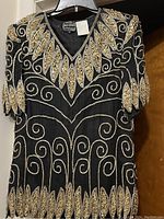 Photo of the black top with gold and black bead embroidery showing leaf and swirl patterns on short sleeves, neckline and hem.