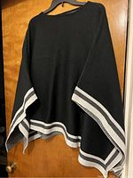 Black knit poncho with light gray and white stripes, hanging on hanger against wood door