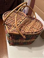 Front view of a rectangular wicker basket with woven pink, blue, and brown fibers, showing the handle and lid details.