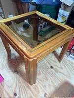 Side angle showing wooden frame and square dark transparent glass top of one end table on wooden floor.