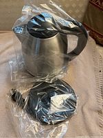 Photo of the Brentwood cordless electric kettle wrapped in plastic, showing stainless steel body, black handle, and power base with cord.