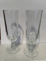 Two tall flute or goblet glasses with decorative bases, clear glass, photographed side by side on white background.