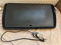 Black electric griddle with cord and temperature control dial shown on fabric surface.