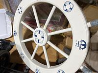 Top view of nautical wooden ship wheel painted white with blue anchor and lifebuoy details, showing eight spokes and rope wrapped rim sections.