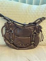 Brown leather tote bag with a front zip pocket, straps with buckle details and drawstring sides shown on a beige fabric background.